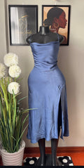 A slate blue satin midi dress with spaghetti straps and a high side slit, displayed on a black mannequin. The mannequin is set in an indoor room with framed wall art, a potted plant with white flowers, and a wooden floor.
