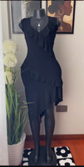 A black sleeveless dress with layered ruffle details and an asymmetrical hem, displayed on a black mannequin in an indoor room setting with framed wall art, a potted plant, and a patterned rug.