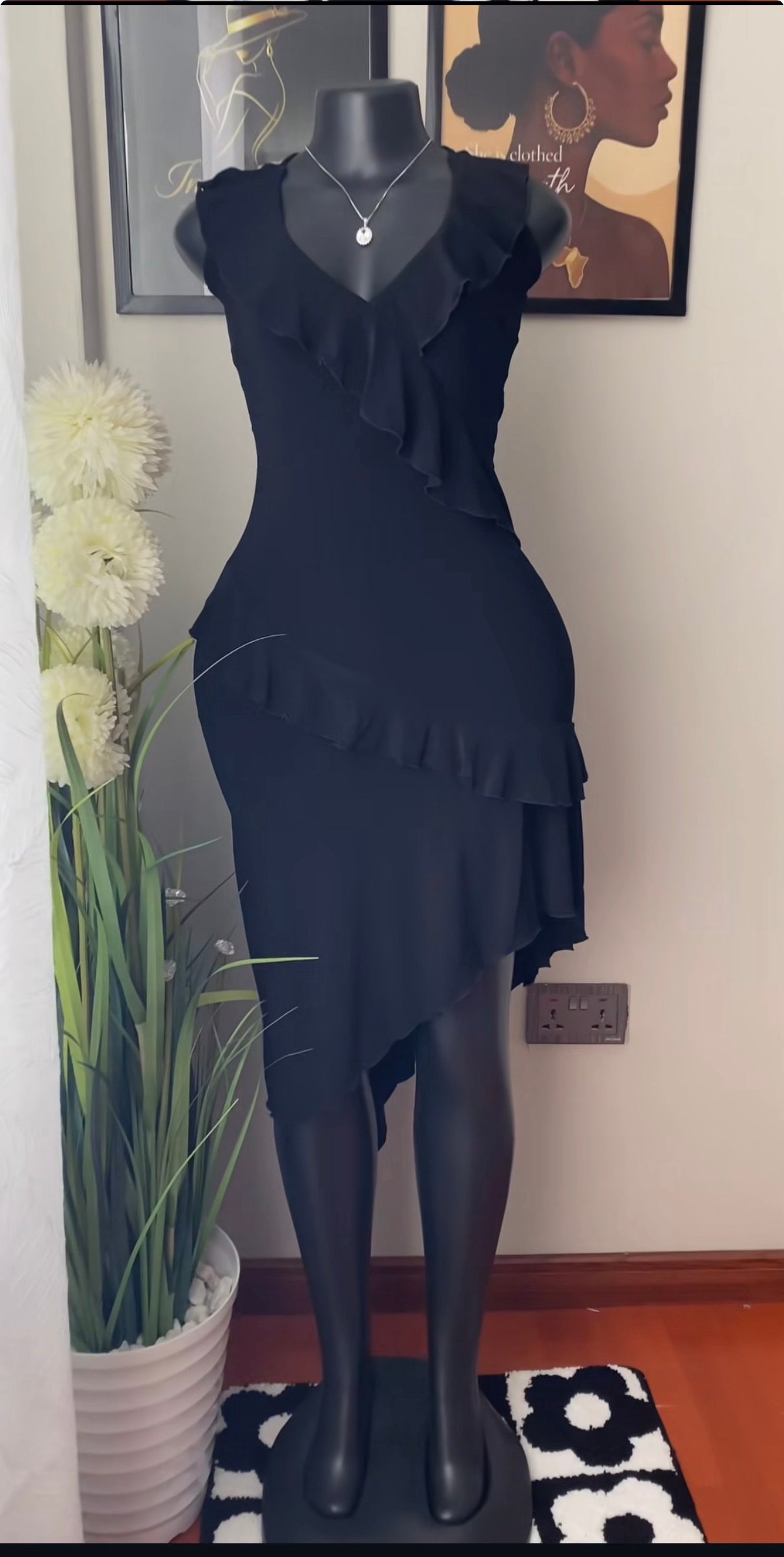 A black sleeveless dress with layered ruffle details and an asymmetrical hem, displayed on a black mannequin in an indoor room setting with framed wall art, a potted plant, and a patterned rug.