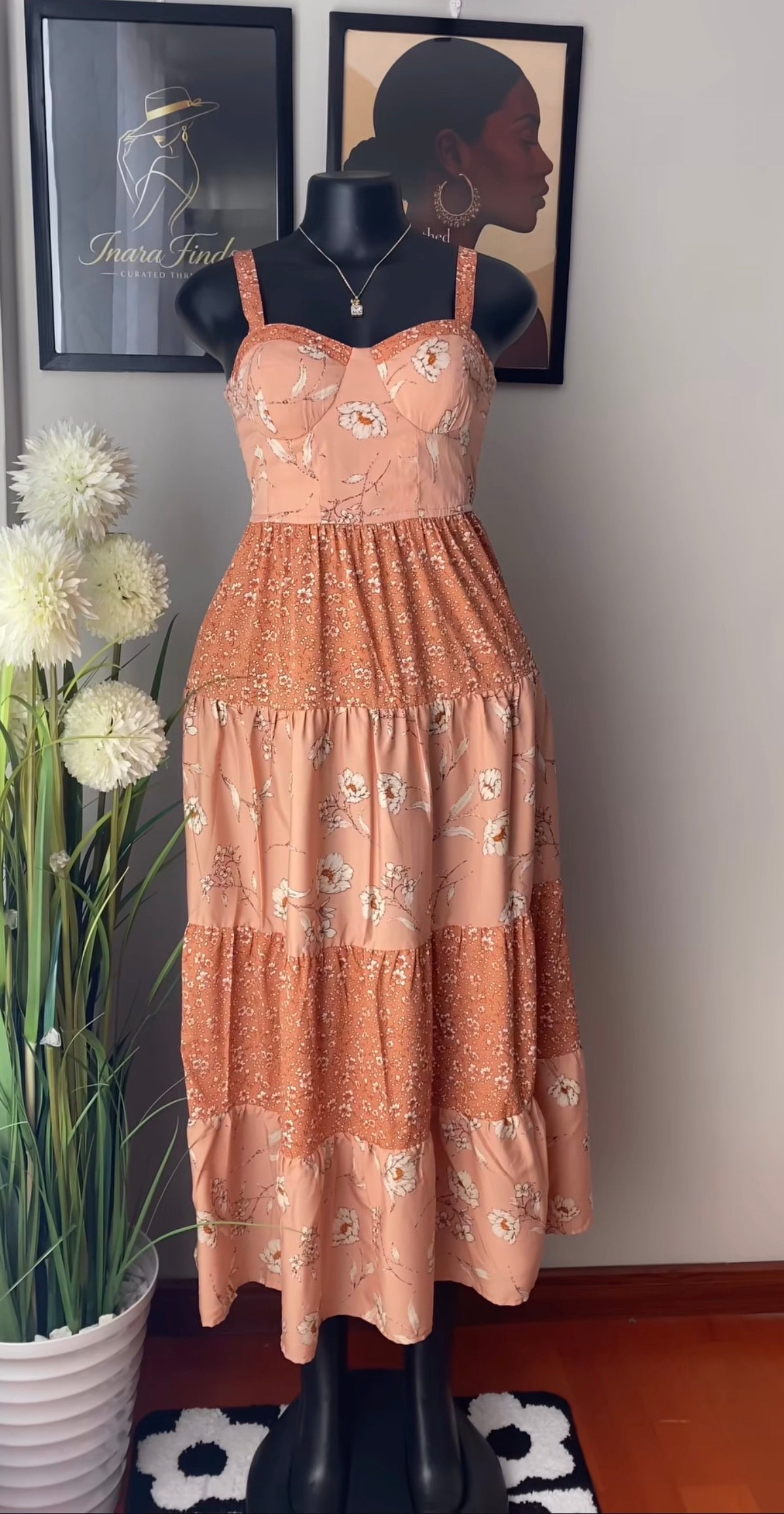 A peach-colored tiered maxi dress with a floral print and embroidered bodice, displayed on a black mannequin. The dress features thin shoulder straps, a sweetheart neckline, and a ruffled tiered skirt.
