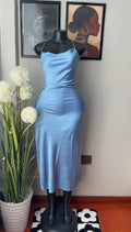 A light blue satin midi dress displayed on a black mannequin torso in an indoor room. The dress has a fitted silhouette with spaghetti straps and a flared skirt.