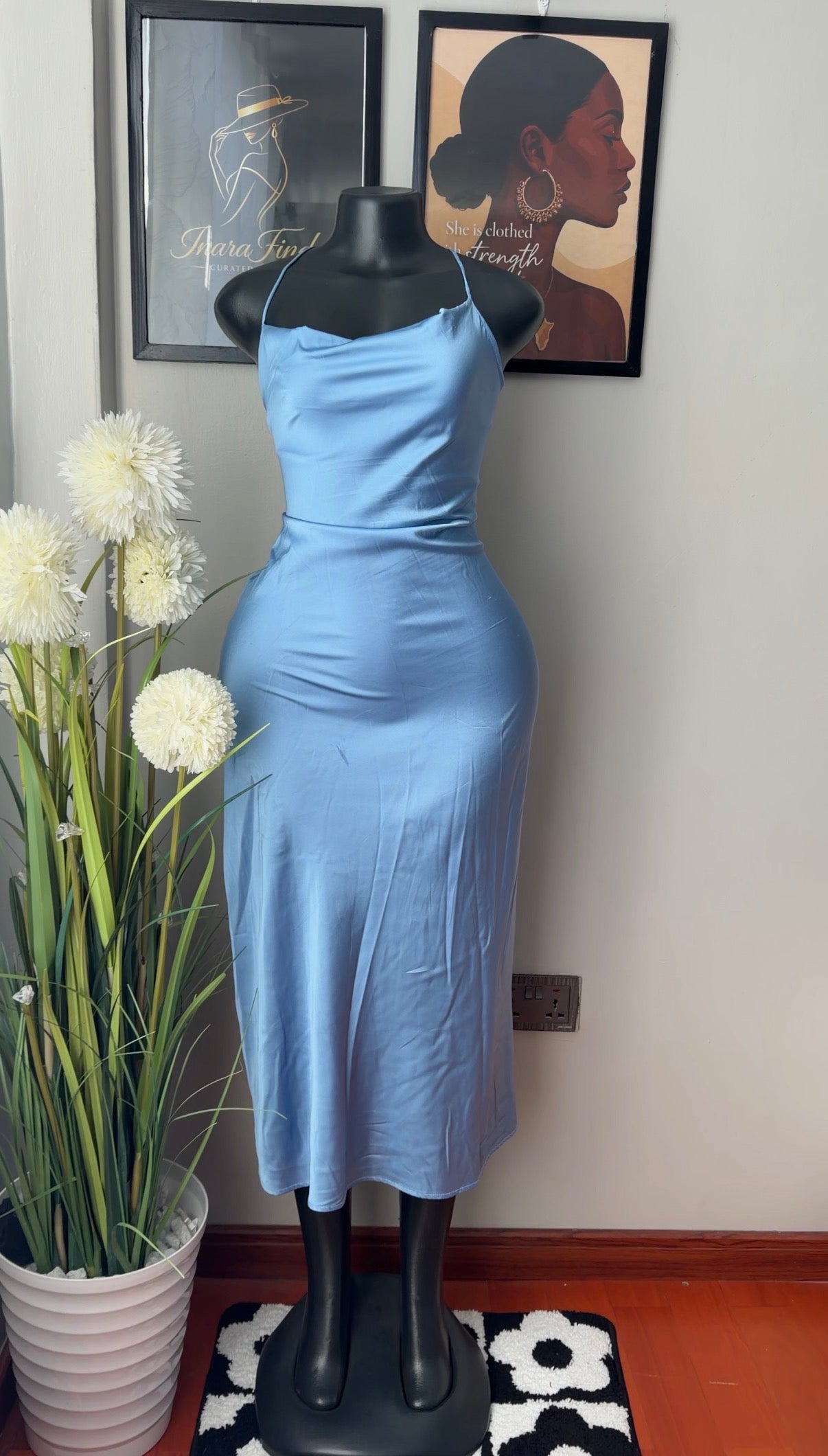 A light blue satin midi dress displayed on a black mannequin torso in an indoor room. The dress has a fitted silhouette with spaghetti straps and a flared skirt.