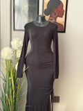 A black long-sleeve bodycon dress displayed on a mannequin torso indoors, with framed wall art and white flowers visible in the background.