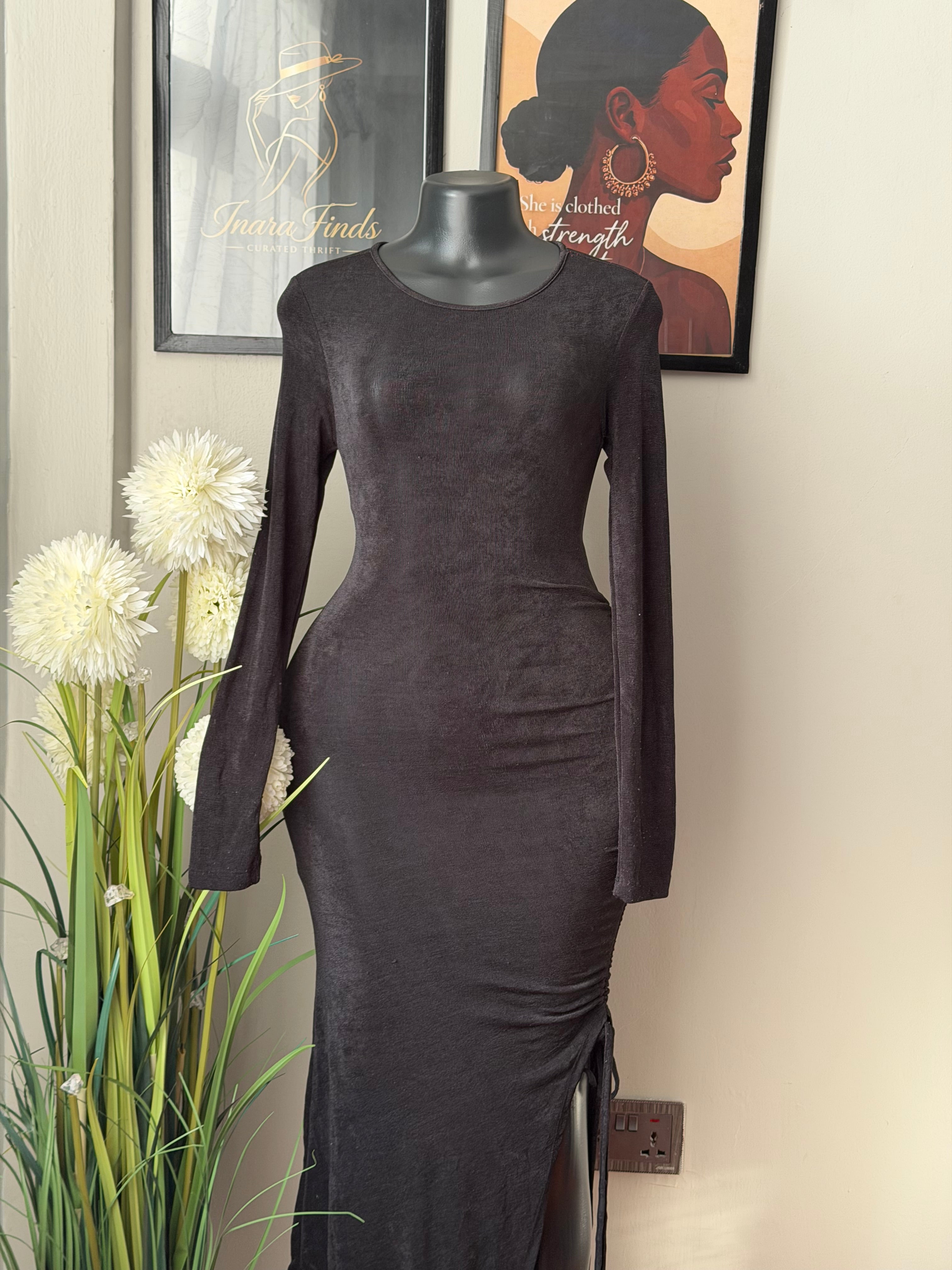 A black long-sleeve bodycon dress displayed on a mannequin torso indoors, with framed wall art and white flowers visible in the background.