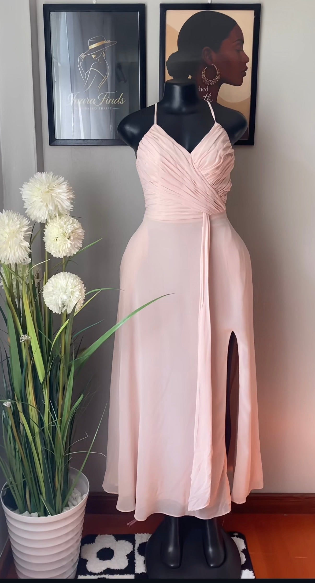A light blush pink sleeveless midi dress with a sweetheart neckline, ruched bodice, and a thigh-high slit, displayed on a black mannequin. The mannequin is set against a gray wall with framed art, and a potted plant with white flowers is on the left.