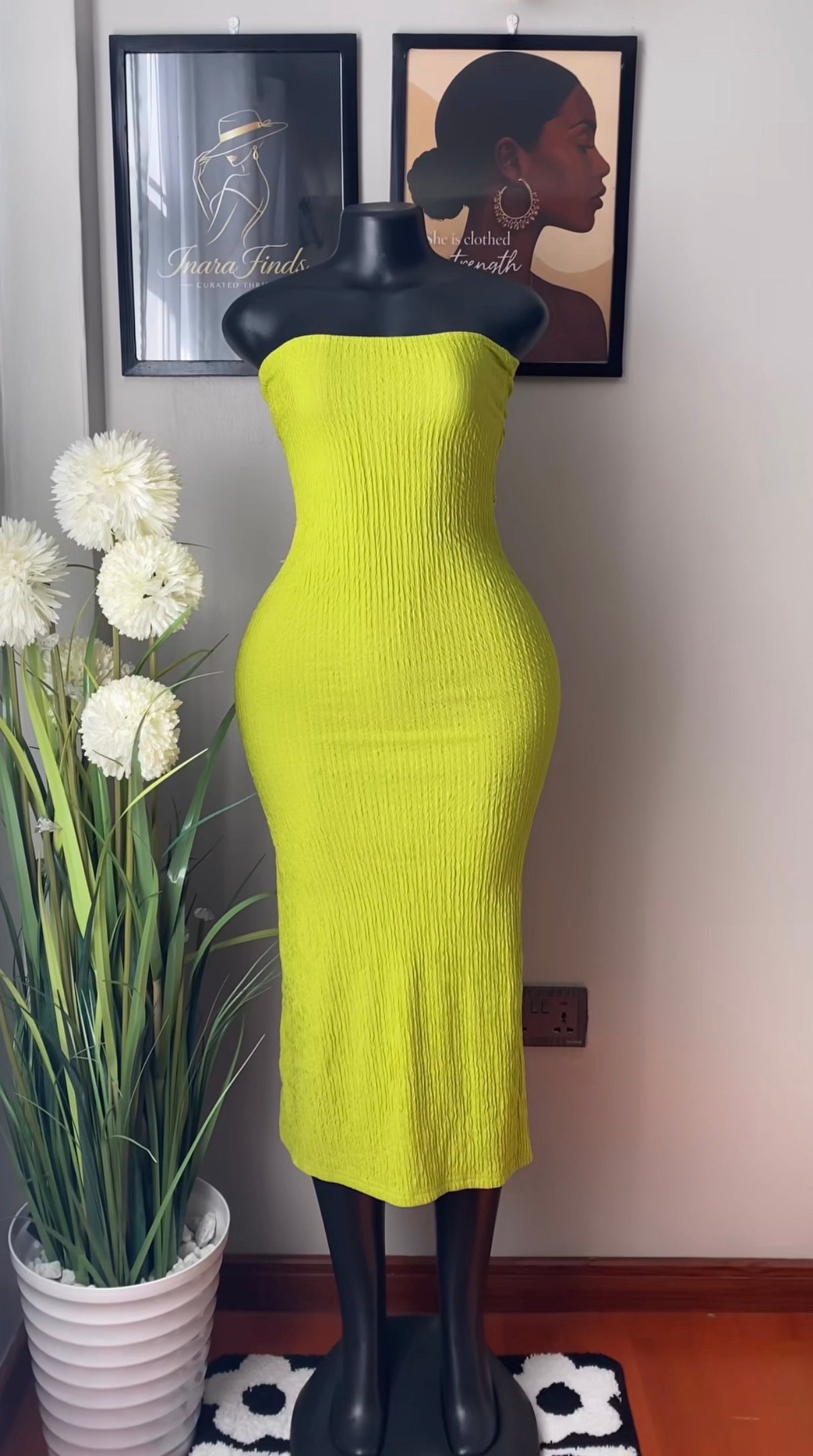 A bright yellow-green ribbed strapless midi dress displayed on a black mannequin, set against a wall with framed art, a potted plant, and a small rug.