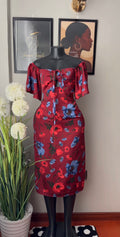A red off-the-shoulder knee-length dress with a blue and red floral print, displayed on a black mannequin. The dress features a gathered waist and short puff sleeves.