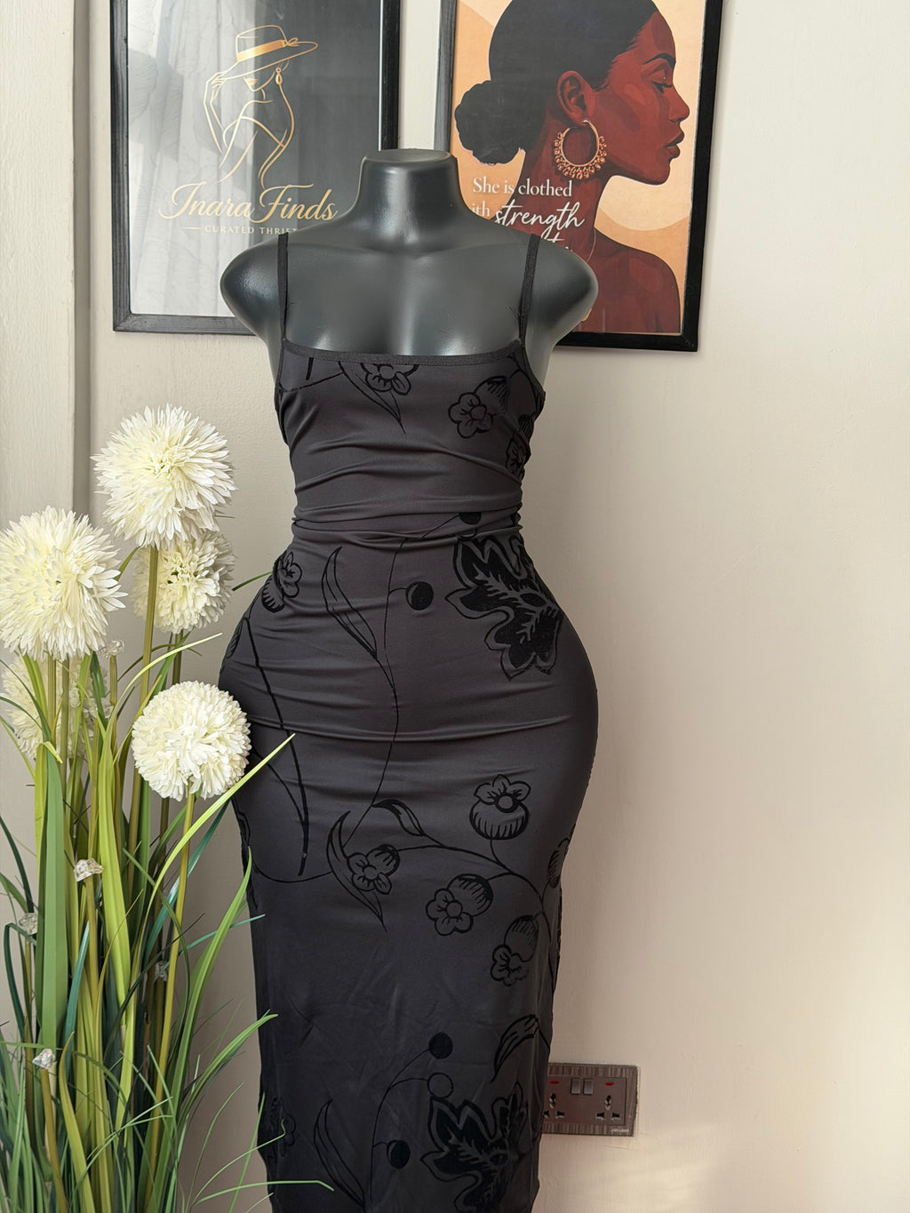 A black ruched midi dress with tonal floral line-art patterns, displayed on a black mannequin torso. The mannequin is set against a neutral wall with framed art and white flowers in the foreground.