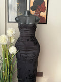 A black ruched midi dress with tonal floral line-art patterns, displayed on a black mannequin torso. The mannequin is set against a neutral wall with framed art and white flowers in the foreground.