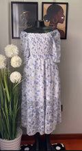 A white off-the-shoulder midi dress with a blue floral print, displayed on a black mannequin. The dress features a smocked bodice and tiered skirt.