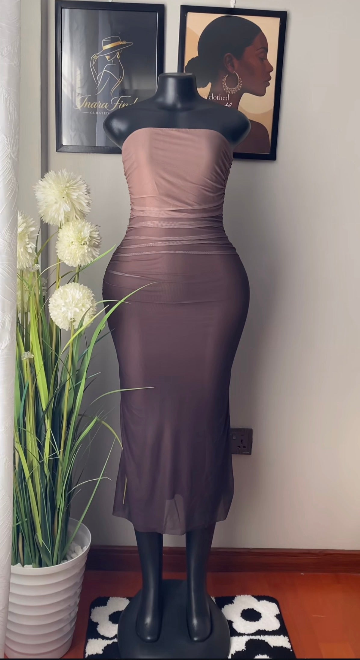 A strapless ruched midi dress in a muted taupe-brown color, displayed on a black mannequin. The dress features a gathered waist and a sheer overlay at the hem.