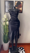 A black satin slip dress displayed on a black mannequin in an indoor room setting, next to a potted plant with white flowers. The dress features a sweetheart neckline, ruched detailing, and a draped side slit.
