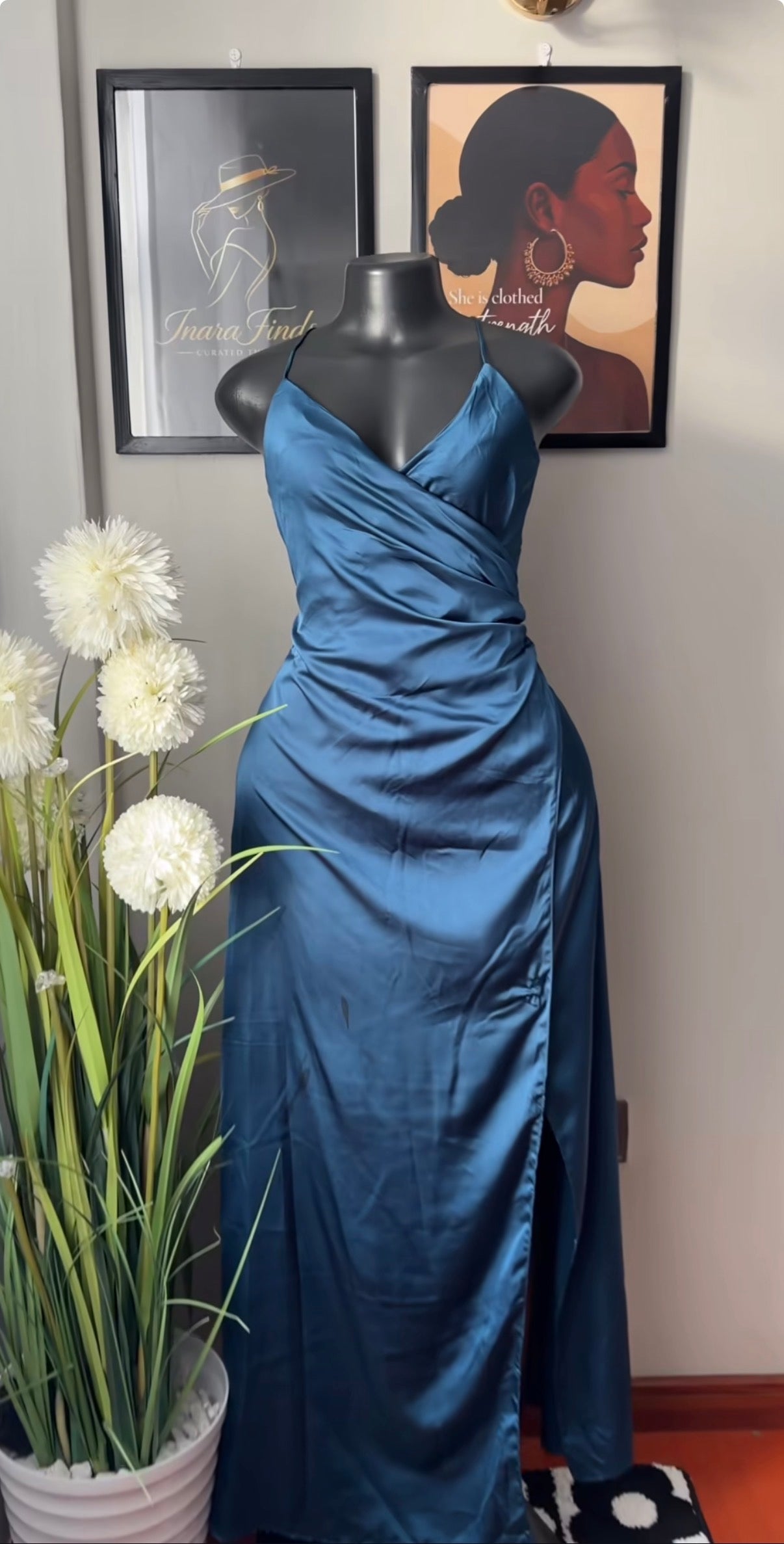 A floor-length blue satin gown displayed on a black mannequin indoors, with framed wall art and a potted plant visible in the background.