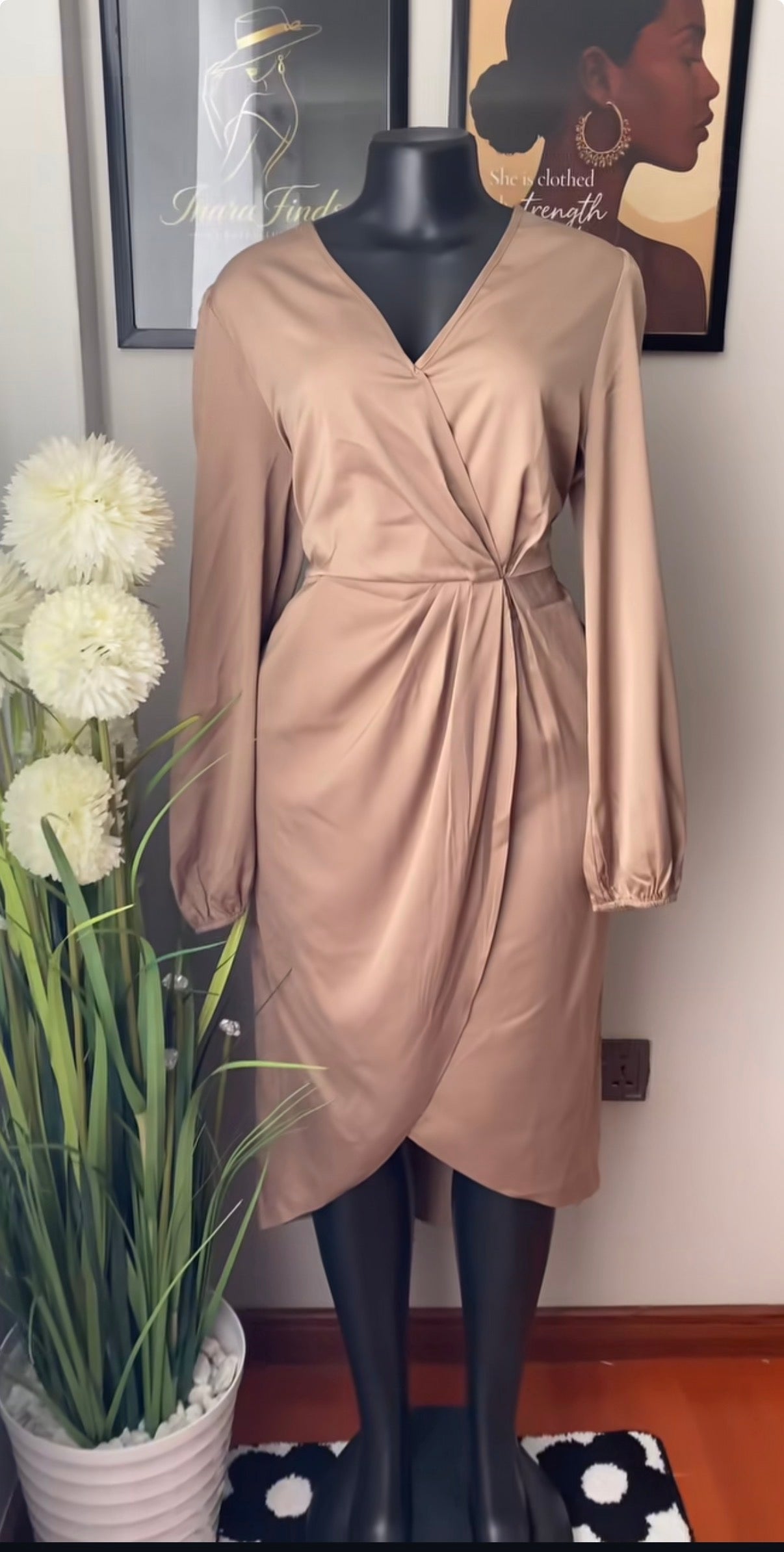 A taupe-beige long-sleeve wrap dress displayed on a black mannequin, set against a home interior background with a potted plant on the left and framed wall art above.
