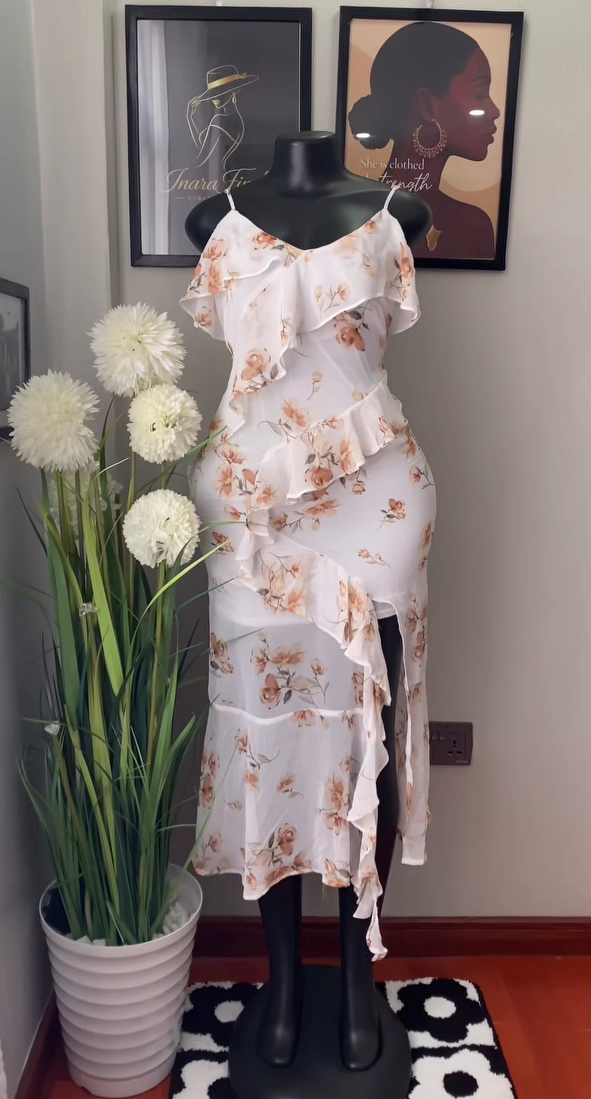 A white ruffled midi dress with a beige and brown floral print, displayed on a black mannequin in an indoor setting with a potted plant and framed wall art.