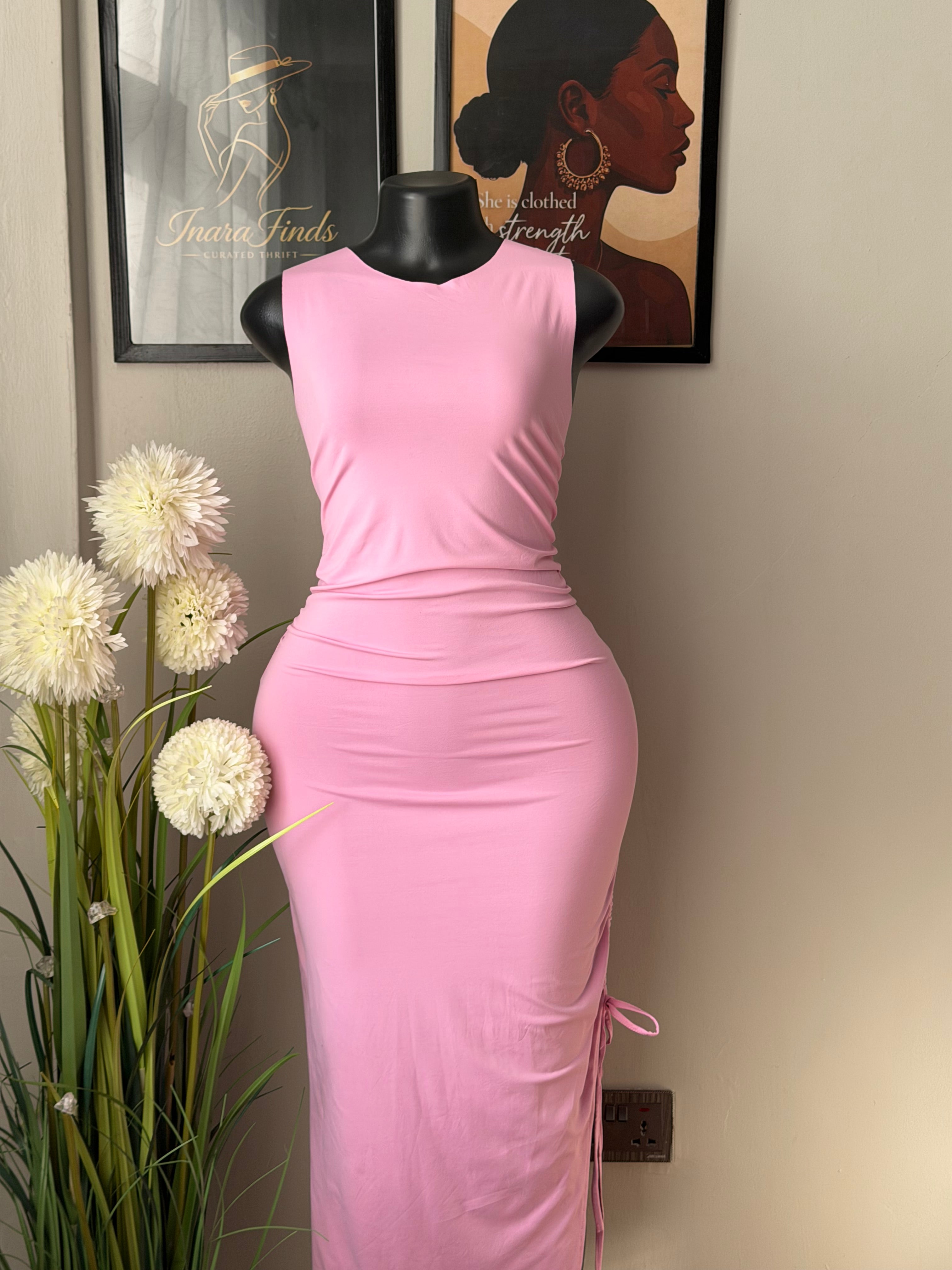 A pink sleeveless fitted dress displayed on a black dress form, photographed indoors against a light wall with framed wall art and white flowers in the foreground.