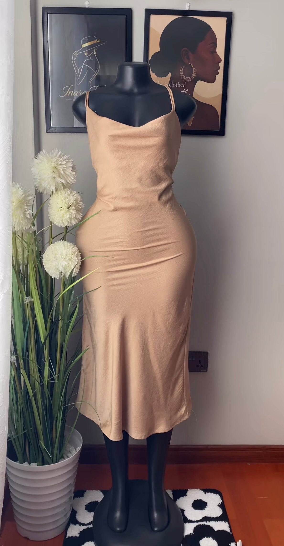 A beige satin slip dress displayed on a black mannequin, set against a wall with framed art and a potted plant.