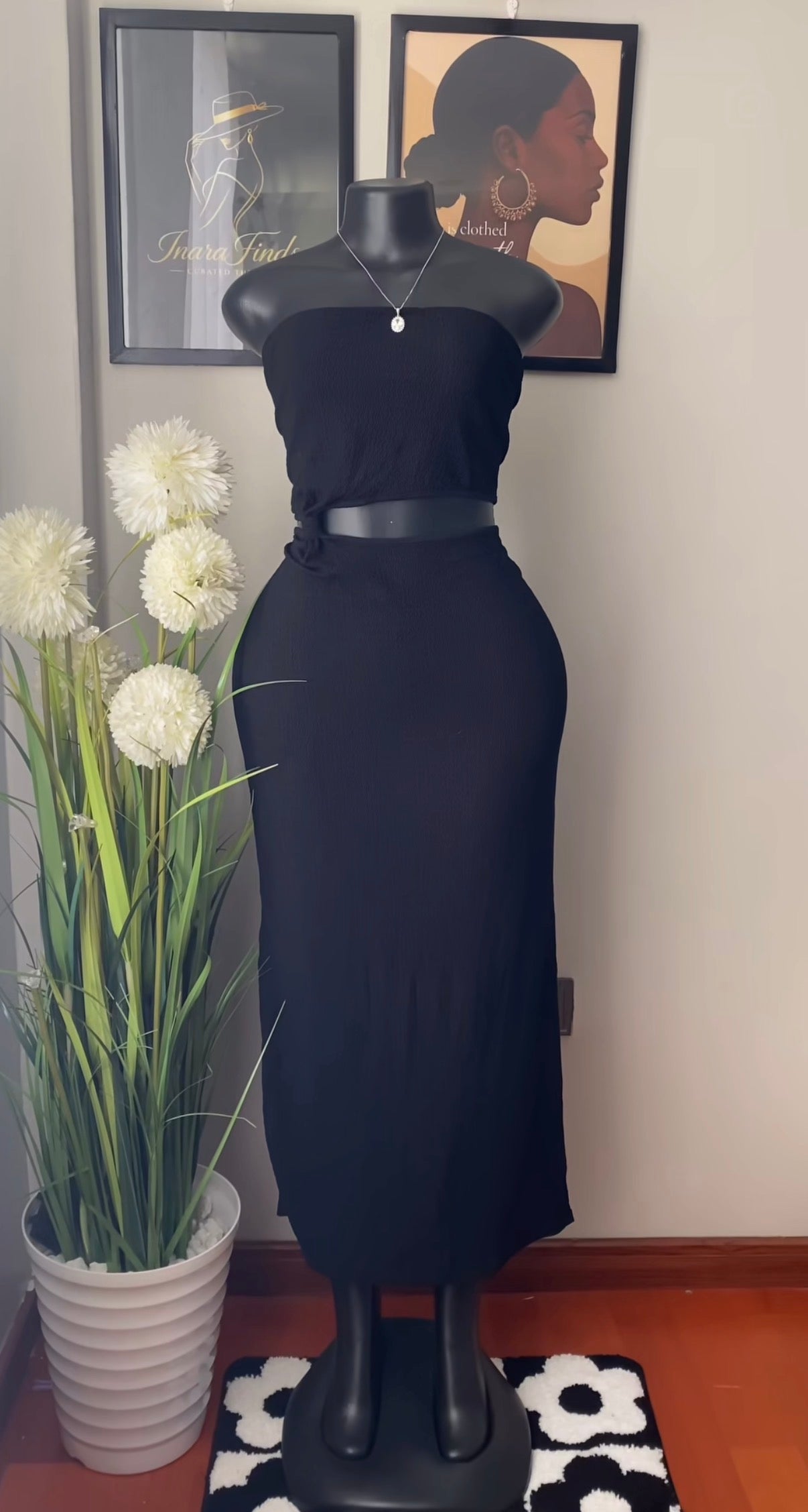 A black strapless midi dress displayed on a black mannequin, styled with a matching black belt. The mannequin is set in an indoor room with framed wall art, a potted plant with white flowers, and a black-and-white patterned rug.