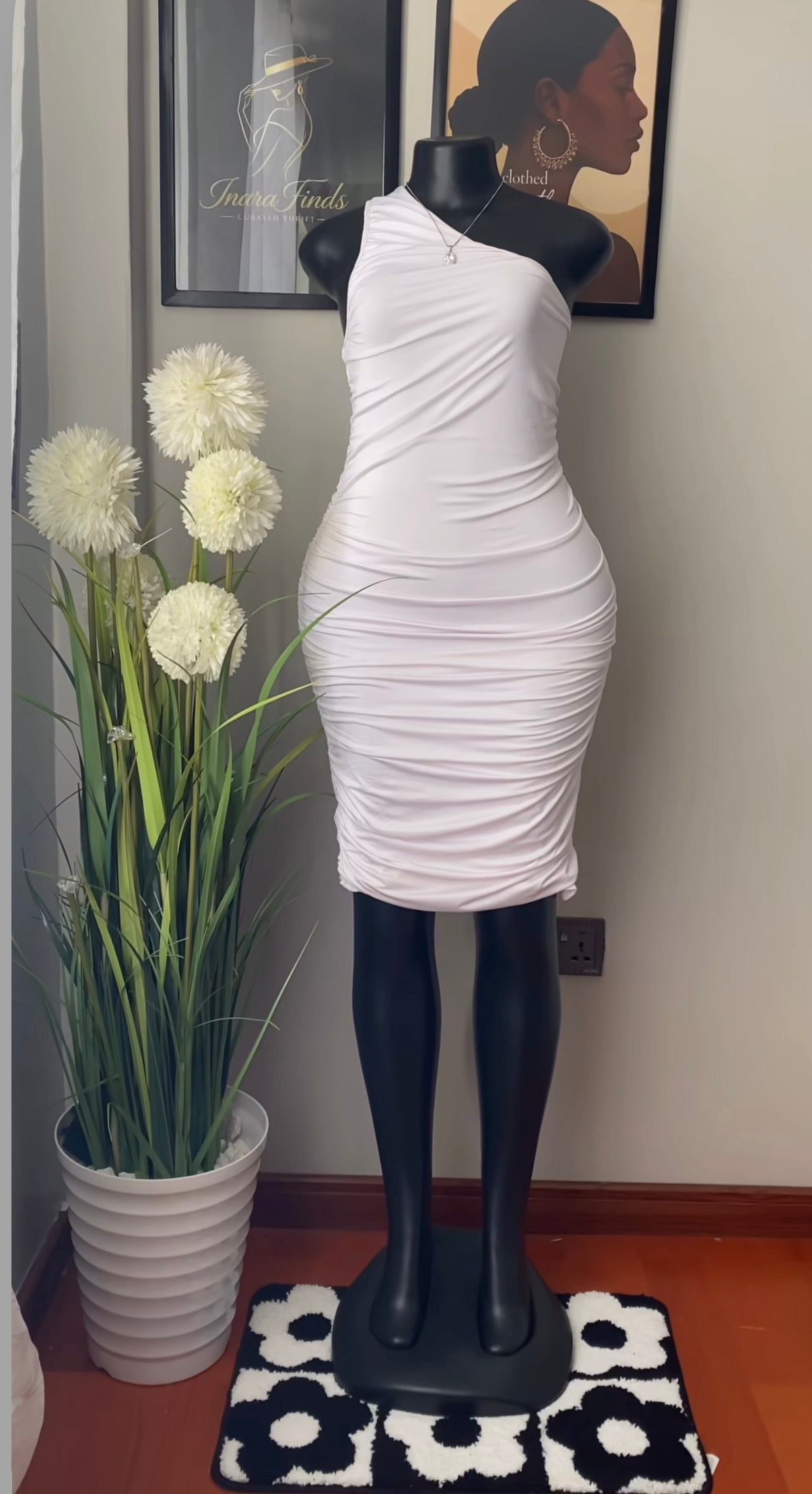 A white ruched one-shoulder bodycon mini dress displayed on a black mannequin indoors, next to a potted plant with white flowers.