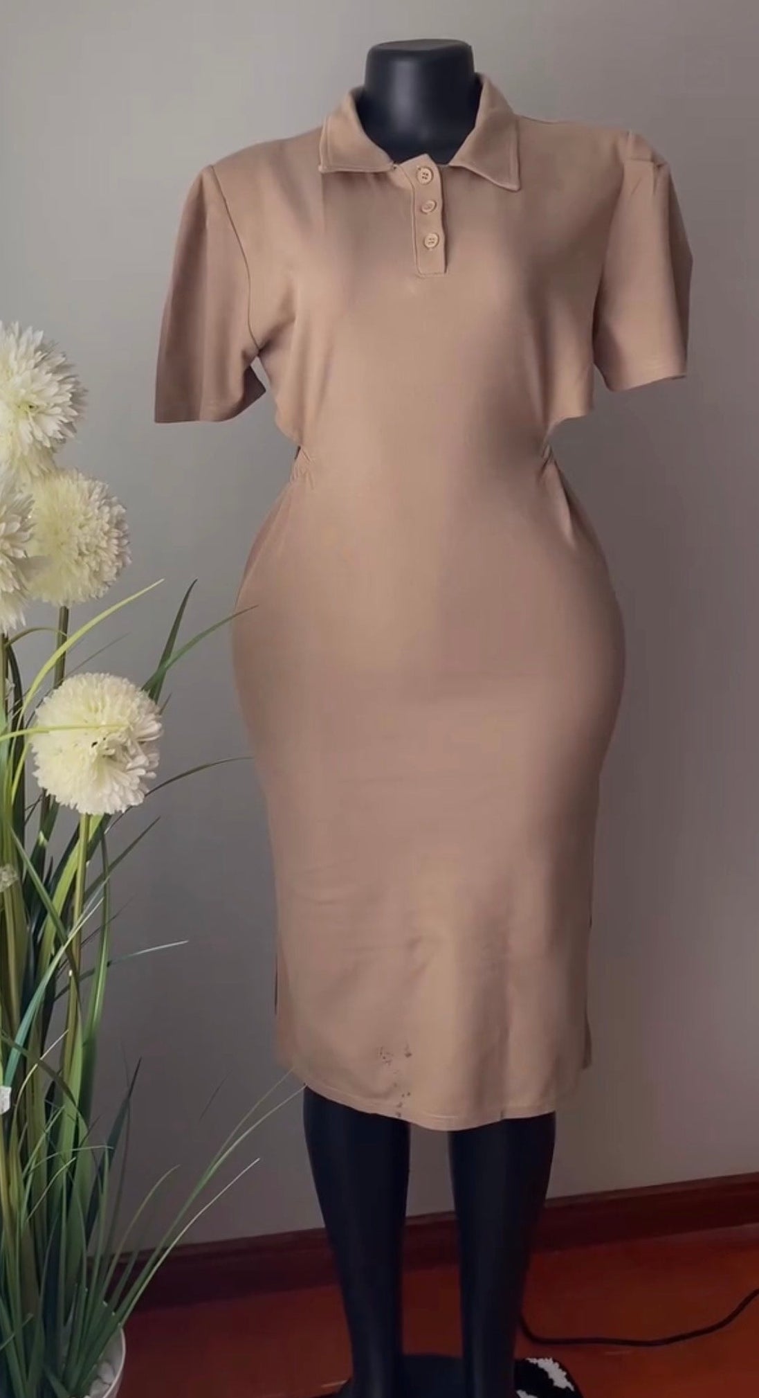 A beige short-sleeve polo dress displayed on a black mannequin, set against a gray wall with a potted plant on the left.