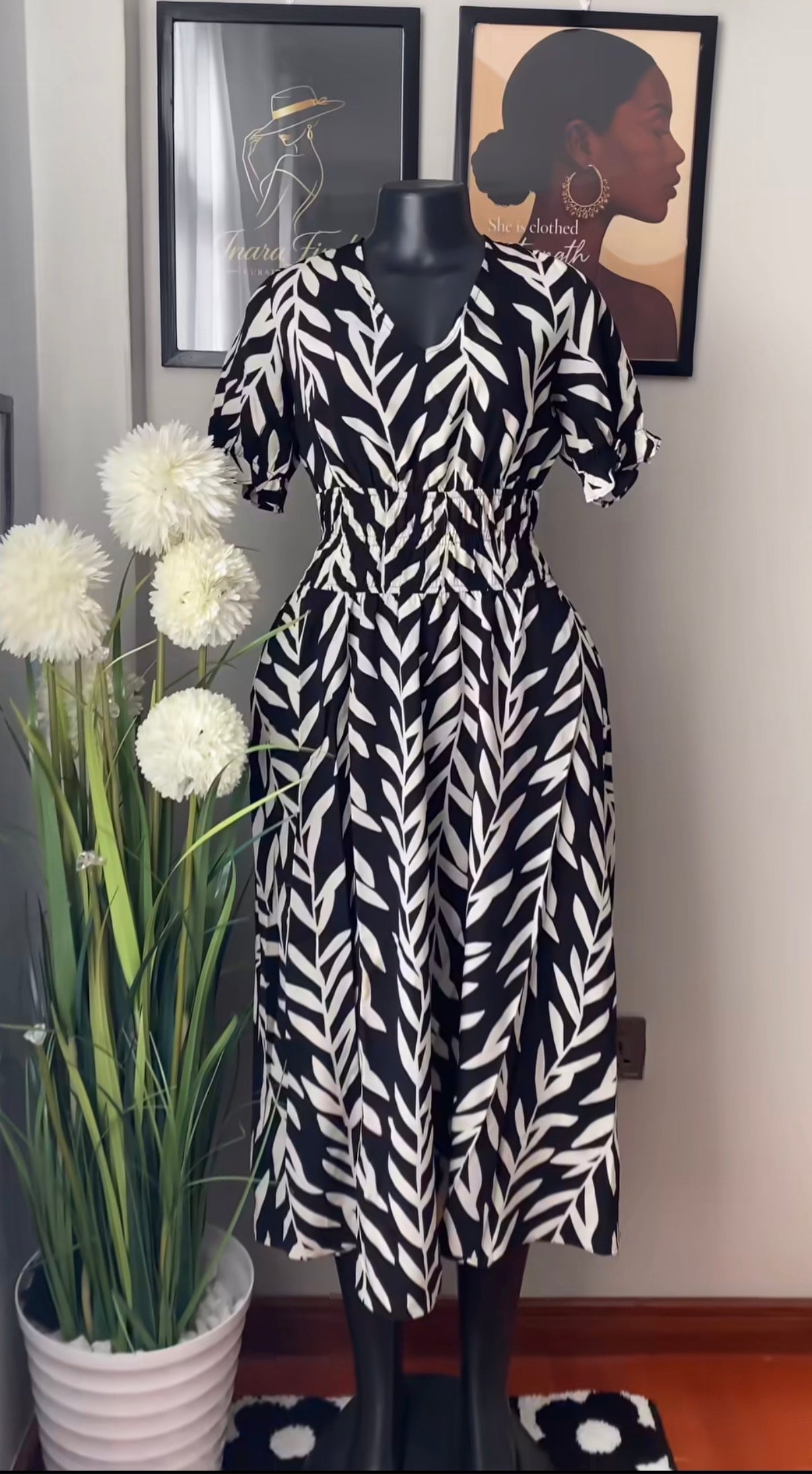 A black and white patterned midi dress with puff sleeves and a V-neckline, displayed on a black mannequin in an indoor setting with a potted plant and framed wall art.