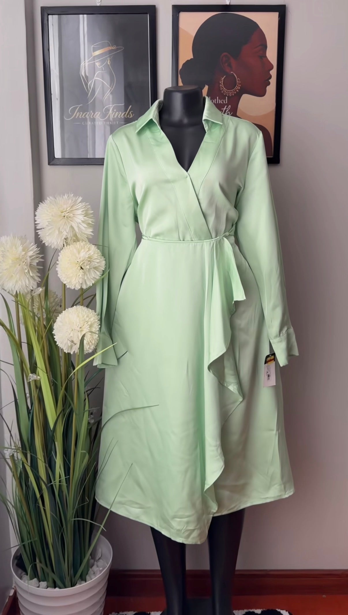 A light mint green wrap dress displayed on a black mannequin, set against a neutral wall with framed art and a potted plant.