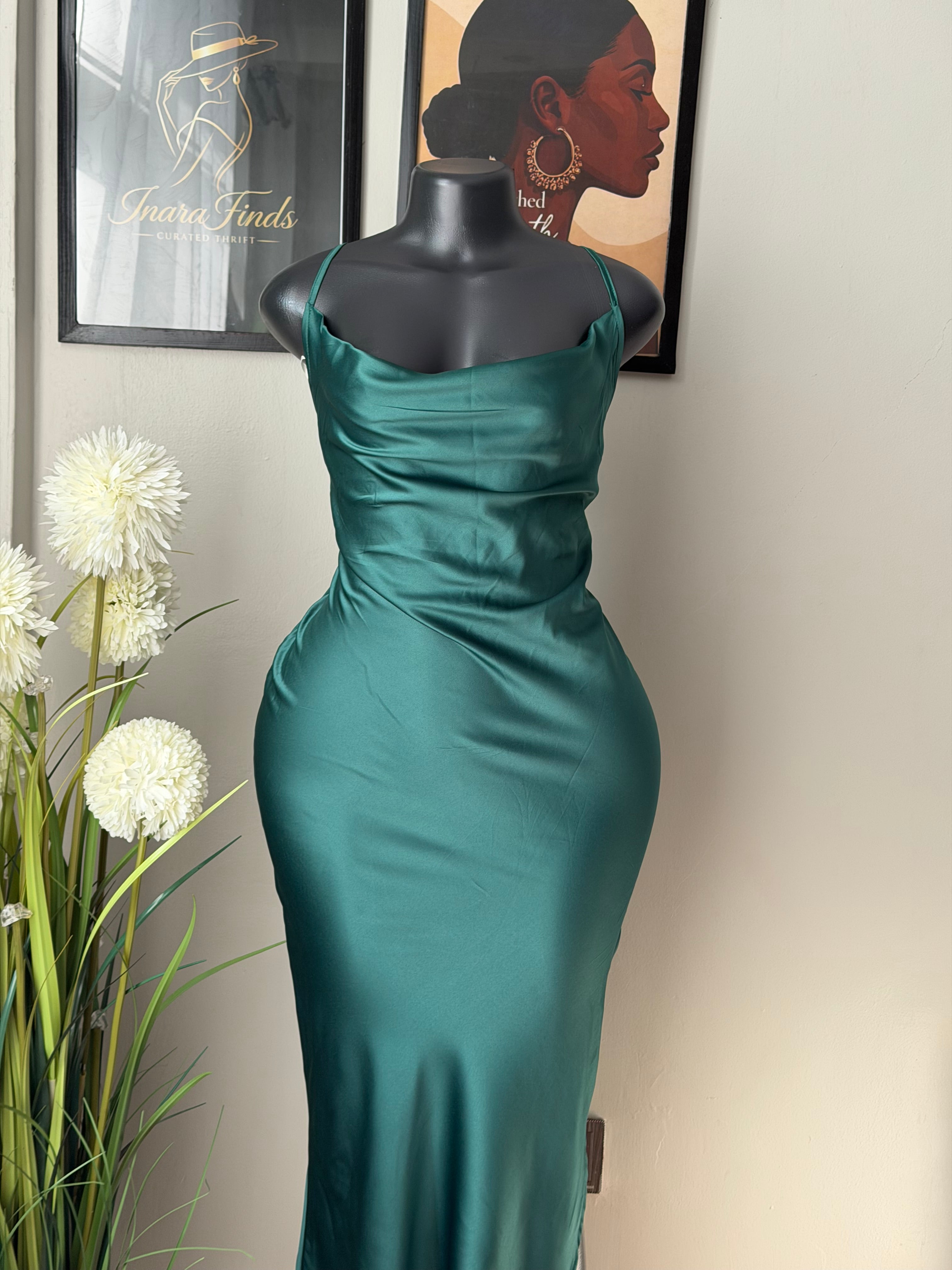 An emerald satin dress displayed on a black mannequin torso, photographed indoors against a light beige wall with framed art and white flowers in the foreground.