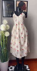A sleeveless cream-colored midi dress with a red and yellow floral print, displayed on a black mannequin. The dress features thin spaghetti straps and a V-neckline.