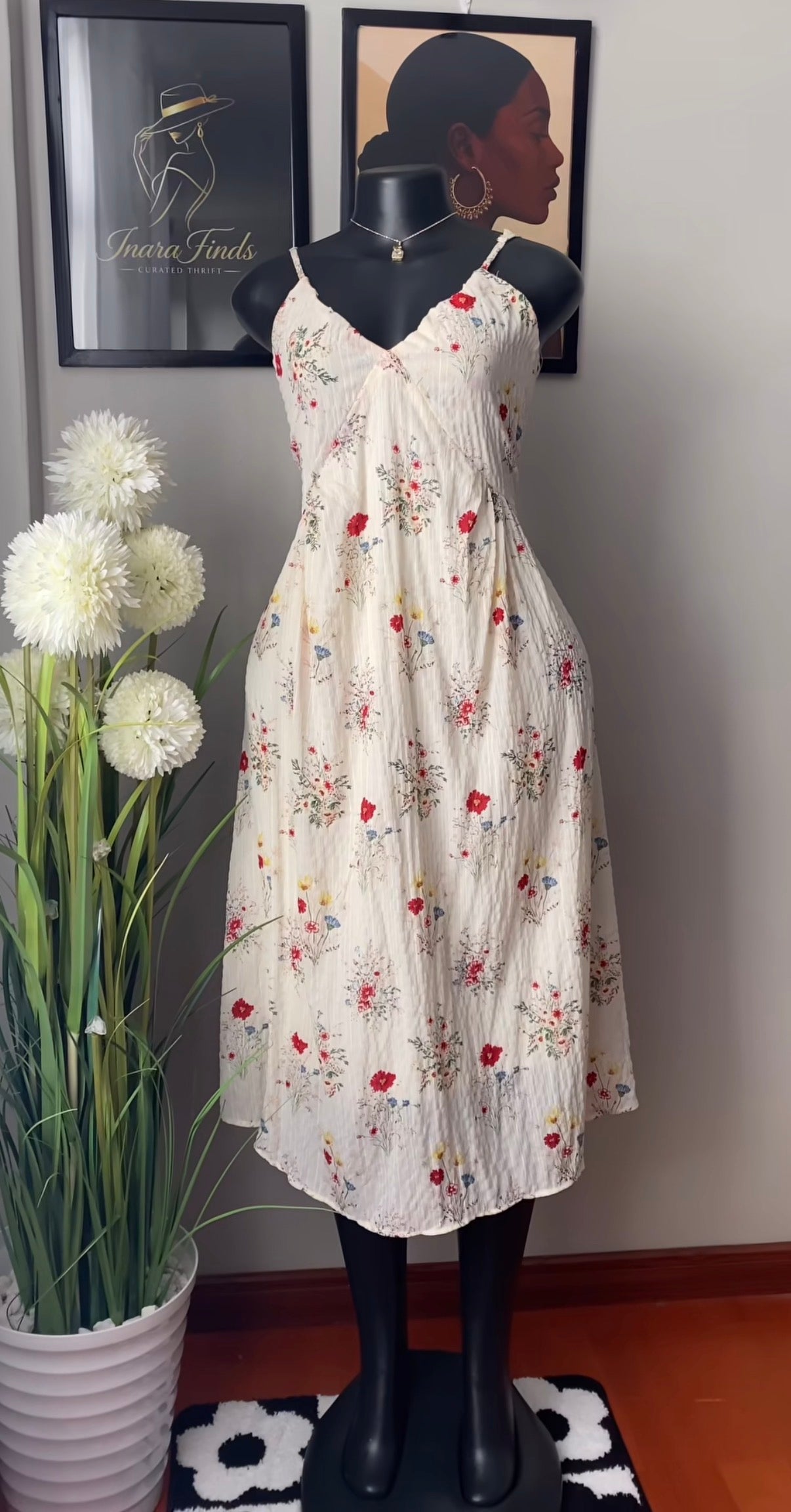 A sleeveless cream-colored midi dress with a red and yellow floral print, displayed on a black mannequin. The dress features thin spaghetti straps and a V-neckline.