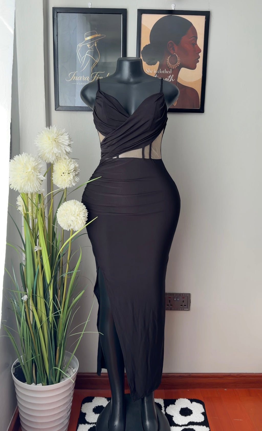 A black sleeveless bodycon maxi dress displayed on a black mannequin, set against a light gray wall with framed art and a potted plant.