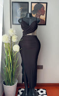 A black sleeveless bodycon maxi dress displayed on a black mannequin, set against a light gray wall with framed art and a potted plant.