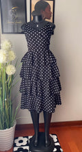 A black sleeveless dress with white polka dots displayed on a black mannequin. The dress features a tiered ruffle skirt and is shown indoors against a beige wall with framed art and a potted plant.