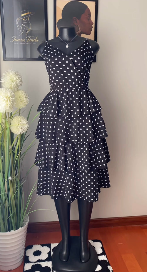 A black sleeveless dress with white polka dots displayed on a black mannequin. The dress features a tiered ruffle skirt and is shown indoors against a beige wall with framed art and a potted plant.