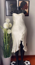 An ivory satin slip dress displayed on a black mannequin, set against a wall with framed art and a potted plant.