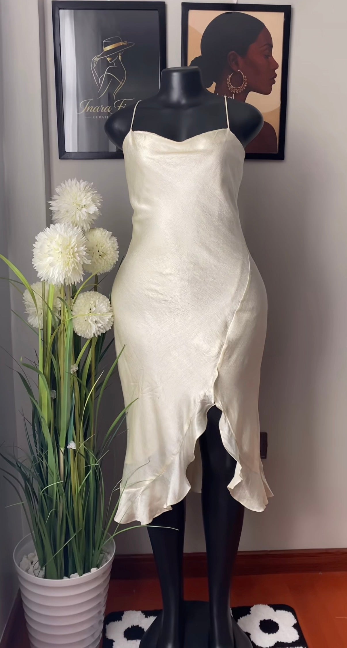 An ivory satin slip dress displayed on a black mannequin, set against a wall with framed art and a potted plant.