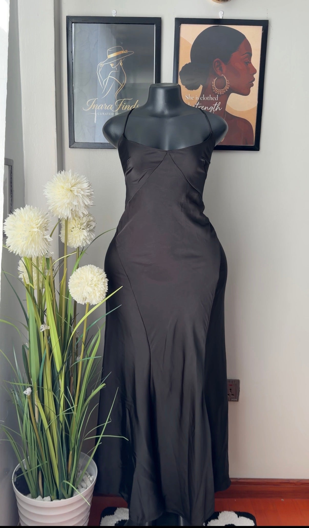 A black satin slip dress displayed on a black mannequin torso in an indoor setting, with framed wall art and a potted plant in the background.
