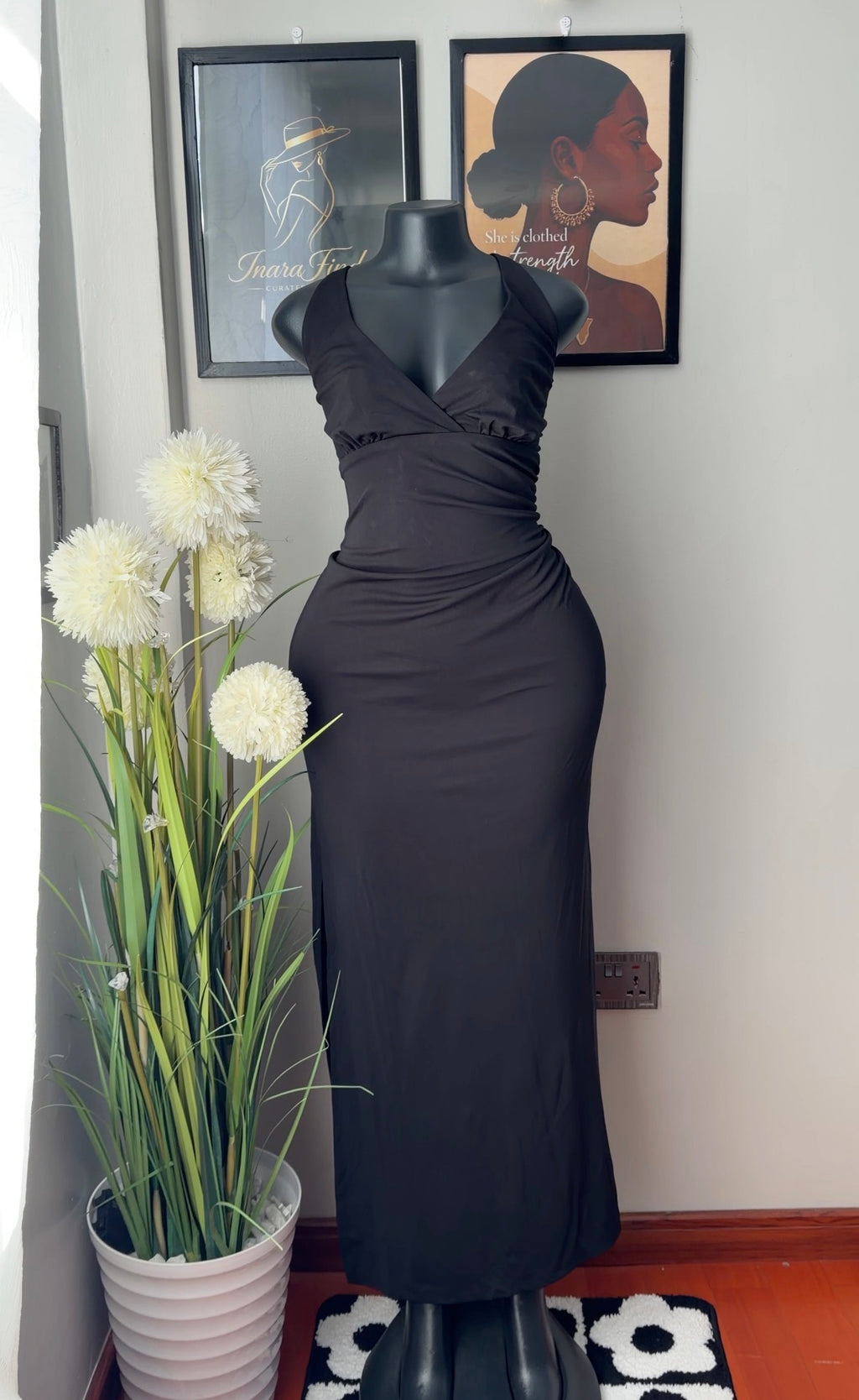A black sleeveless wrap-style maxi dress displayed on a black mannequin indoors, with framed wall art and a potted plant in the background.