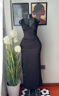 A black sleeveless wrap-style maxi dress displayed on a black mannequin indoors, with framed wall art and a potted plant in the background.