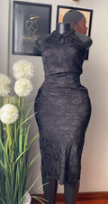 A black lace sleeveless dress displayed on a black mannequin indoors, with framed wall art and a potted plant visible in the background.