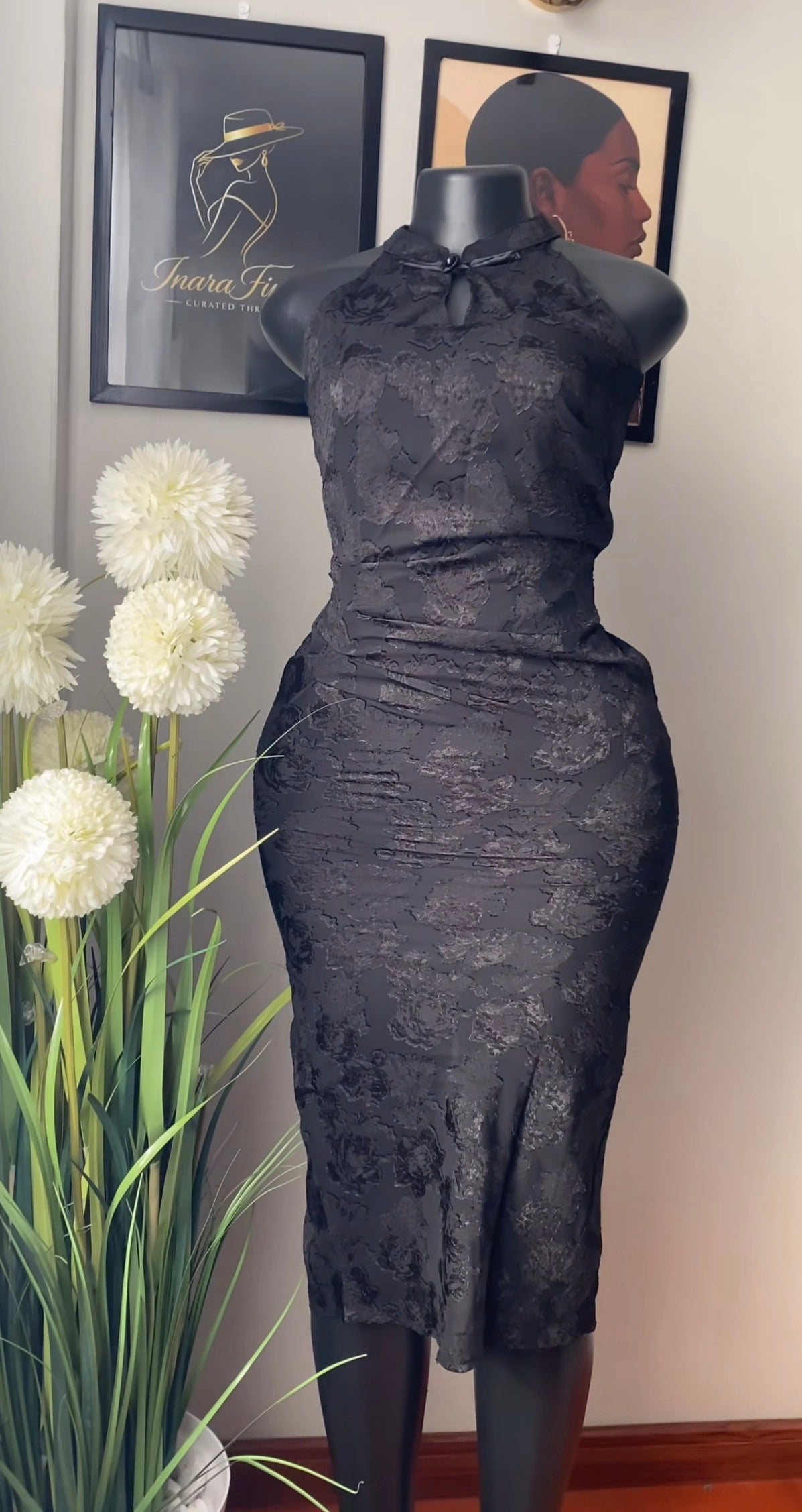 A black lace sleeveless dress displayed on a black mannequin indoors, with framed wall art and a potted plant visible in the background.
