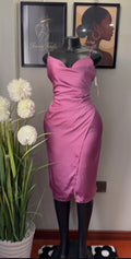 A mauve pink satin midi dress displayed on a black mannequin torso, set against a wall with framed art, a wall lamp, and a potted plant.
