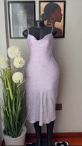 A light lavender spaghetti-strap midi slip dress displayed on a black mannequin torso, set against a wall with framed art and a potted plant.