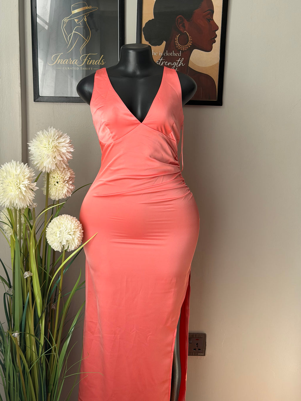 A coral pink sleeveless dress displayed on a black mannequin torso, photographed indoors against a light beige wall with framed art and white flowers in the foreground.